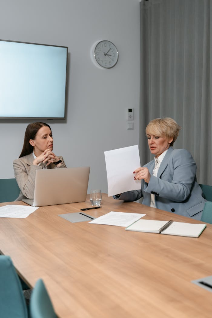 Two women in corporate attire discussing documents in an office meeting room.